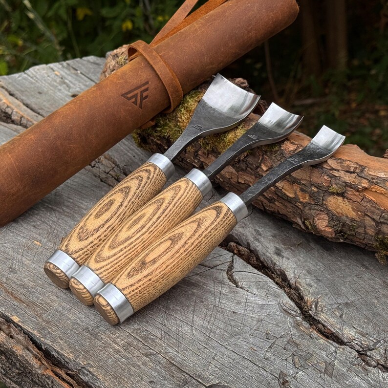 May include: Three wood carving gouges with curved metal blades and light brown wood grain handles, featuring silver metal ferrules. The tools are accompanied by a brown leather roll case embossed with a geometric logo featuring the letters AF.
