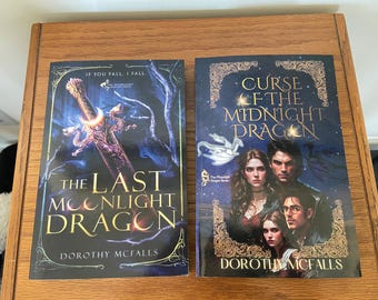 May include: Two fantasy novel covers with a dark blue background. The first cover features a silver sword with a dragon design and the text "The Last Moonlight Dragon" by Dorothy Mcfalls. The second cover features a dragon design and the text "Curse of the Midnight Dragon" by Dorothy Mcfalls.