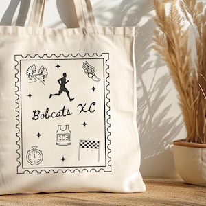 May include: Cream-colored tote bag featuring a black graphic design. The design includes a runner, trees, a stopwatch, a jersey, a checkered flag, and the words "Bobcats XC". The bag has two handles.