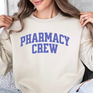Könnte beinhalten: Ein cremefarbener Pullover mit dem Text "Pharmacy Crew" in Blau.