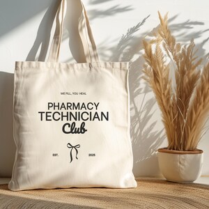 Könnte beinhalten: Eine weiße Canvas-Tragetasche mit schwarzem Text, der "We fill, you heal. Pharmacy Technician Club. Est. 2025." lautet. Die Tasche hat schwarze Träger und eine schwarze Schleife.