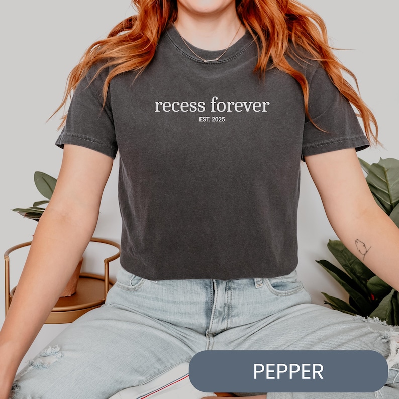 Op de afbeelding: Een donkergrijs gecropped T-shirt met de tekst "recess forever EST. 2025" in witte letters. Het model draagt het shirt met lichtblauwe jeans.