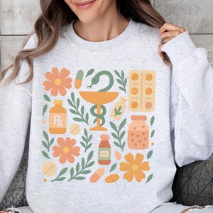 Könnte beinhalten: Ein hellgraues Sweatshirt mit einem farbenfrohen Apotheken-Themen-Design. Das Design umfasst orangefarbene Blumen, grüne Blätter, Pillenflaschen, Kapseln und ein Apotheken-Symbol. Die Gesamtästhetik ist eine fröhliche und einladende Illustration.