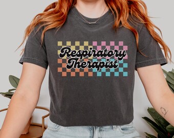 Puede incluir: Una camiseta negra recortada con un patrón de cuadros coloridos y el texto "Respiratory Therapist" en una fuente retro.