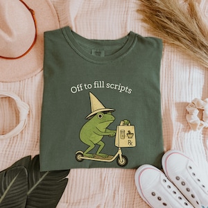 Könnte beinhalten: Grünes T-Shirt mit dem Text "Off to fill scripts". Das Shirt zeigt einen Cartoon-Frosch mit Hut, der einen Roller fährt und eine Apotheken-Tasche hält. Weiße Turnschuhe sind unten rechts zu sehen.
