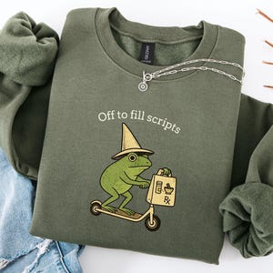 Puede incluir: Sudadera verde oliva con el texto "Off to fill scripts" sobre una rana de dibujos animados montada en un scooter y llevando una bolsa de farmacia. Un collar plateado está drapeado sobre la sudadera. La sudadera está doblada sobre unos vaqueros azules.