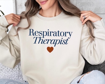 Może przedstawiać: Kremowa bluza z napisem "Respiratory Therapist" w kolorze granatowym i brązowym sercem. Bluza ma swobodny styl, odpowiedni do noszenia na co dzień.