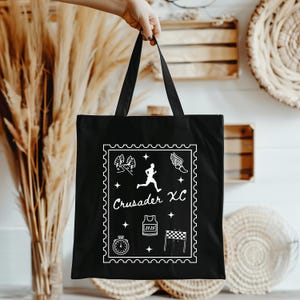 May include: Black tote bag with a white graphic featuring a runner, trees, stars, and the text "Crusader XC". Additional graphics include a stopwatch, jersey, and checkered flag. The bag is held by a hand.