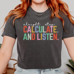 May include: Dark gray t-shirt with the words "alright stop. CALCULATE. AND LISTEN." in colorful block letters. The shirt is a casual style, suitable for everyday wear.