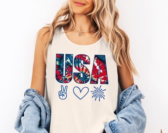 May include: Cream-colored tank top with the word "USA" in tie-dye lettering. Below the text are blue and white peace sign, a blue heart, and a blue firework graphic. The top is sleeveless.