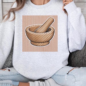 Könnte beinhalten: Hellgraues Sweatshirt mit einer Grafik eines hölzernen Mörsers und Stößels. Der Mörser und Stößel sind braun mit diagonalen Streifen, vor einem Hintergrund aus vertikalen braunen Streifen.