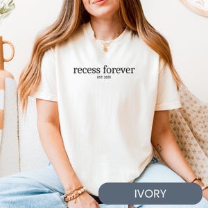 Op de afbeelding: Een wit T-shirt met de tekst "recess forever est. 2025" gedrukt op de voorkant. Het shirt wordt gedragen door een model dat een blauwe jeans draagt. De kleur van het shirt is ivoor.