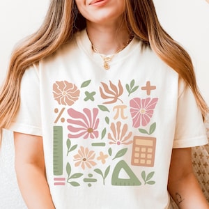 May include: A white t-shirt with a graphic design featuring a variety of math symbols and flowers in pink, green, and brown colors. The symbols include a plus sign, a minus sign, a multiplication sign, a division sign, a percentage sign, a pi symbol, and an equal sign. The flowers are stylized and have a simple design.