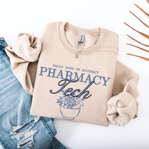 Könnte beinhalten: Beiges Sweatshirt mit dem Text "DAILY DOSE OF ACCURACY PHARMACY Tech" in Blau und schwarzer Schrift. Eine Mörser- und Stößel-Grafik mit Blumen befindet sich unter dem Text. Eine silberne Halskette liegt auf dem Sweatshirt.