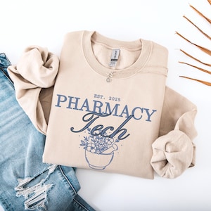 Könnte beinhalten: Beiges Sweatshirt mit dem Text "PHARMACY Tech" in Blau und schwarzer Schrift, darüber "EST. 2025". Eine Mörser- und Stößel-Grafik mit Blumen befindet sich unter dem Text. Eine silberne Halskette liegt über dem Sweatshirt.
