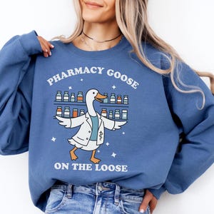Könnte beinhalten: Ein blaues Sweatshirt mit dem Text "PHARMACY GOOSE ON THE LOOSE". Die Grafik zeigt eine Gans mit einem Laborkittel vor Regalen mit Medikamentenflaschen. Die Gans hat ihre Flügel ausgebreitet. Das Sweatshirt hat eine einfarbige blaue Farbe.