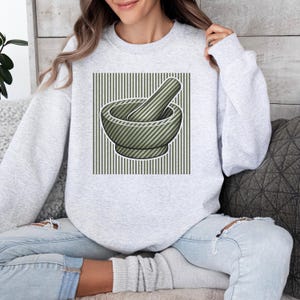 Könnte beinhalten: Ein hellgraues Sweatshirt mit einem grünen Mörser und Stößel-Motiv. Der Mörser und Stößel sind gestreift und vor einem gestreiften Hintergrund platziert. Das Sweatshirt ist ein lässiger, bequemer Stil.