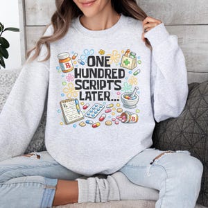 Könnte beinhalten: Ein hellgraues Sweatshirt mit einem farbenfrohen Grafikdesign mit verschreibungspflichtigen Pillen, einem Mörser und Stößel sowie dem Text "ONE HUNDRED SCRIPTS LATER...". Das Design umfasst eine Rezeptflasche und einen Notizblock.