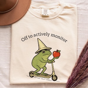 May include: Cream-colored t-shirt featuring a green frog wearing a hat and riding a scooter while holding a red apple. The text "Off to actively monitor" is printed above the frog.
