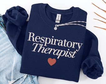 Może przedstawiać: Granatowa bluza z napisem "Respiratory Therapist" w kolorze białym, czerwonym sercem i srebrnym naszyjnikiem. Bluza jest złożona na niebieskich dżinsach.