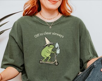 Pode incluir: Camiseta verde com a frase "Off to clear airways". O design apresenta um sapo de desenho animado com um chapéu, andando de scooter e segurando um inalador. O sapo é verde e a scooter é bege.
