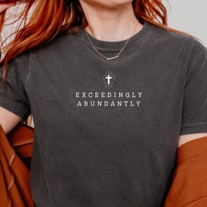 May include: A dark gray t-shirt with a white cross and the text "Exceedingly Abundantly" printed on the front.