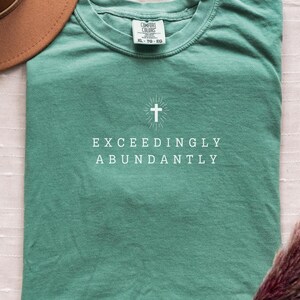 May include: A teal green t-shirt with a white cross and the text "Exceedingly Abundantly" printed on the front.