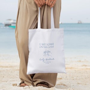 May include: White tote bag with blue text that reads "Last Toast on the Coast" and a blue illustration of two hands holding up champagne glasses. The text "Emily's Bachelorette The Hamptons 1.1.2024" is also printed on the bag.