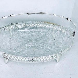 May include: Oval serving tray with a clear, patterned glass insert and a white metal frame. The tray has a handle and sits on three small feet. The glass insert features a starburst design.