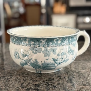 Op de afbeelding: Antieke keramische kom met een wit en blauw bloemenpatroon. De kom heeft een gedetailleerd bloemenpatroon in blauw- en wittinten, met een decoratieve rand rond de rand. De kom heeft een handvat.