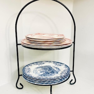 May include: A two-tiered black metal serving stand with stacks of vintage plates. The top tier has pink floral plates, the bottom tier has blue and white scenic plates. The stand has a curved handle. The plates are ideal for serving cakes.