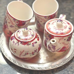 Könnte beinhalten: Ein weißes Keramik-Teeservice mit floralen und textlichen Details, darunter eine Teekanne, eine Zuckerdose und zwei Tassen, angeordnet auf einem silbernen Tablett. Das Set zeigt rote und lila Blumenmuster und die Worte "Enjoy the joy, laugh, smiles, and friends."
