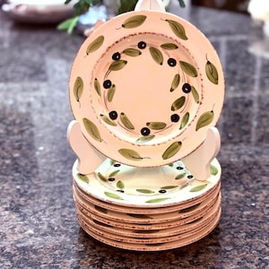 Könnte beinhalten: Ein Stapel cremefarbener Keramikteller mit handgemaltem Olivenzweig-Design. Jeder Teller hat einen braunen Rand und ein zentrales Oliven- und Blattmotiv. Die Teller sind auf einem weißen Tellerständer gestapelt.