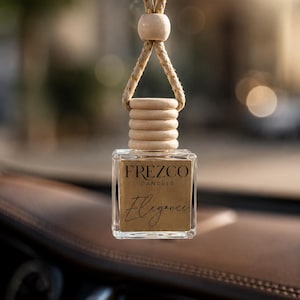 May include: A square glass car air freshener bottle with a light wood cap and a braided beige cord. The bottle features a tan label with the black text FREZCO CANDLES and the word Elegance in a script font, hanging from a rearview mirror.