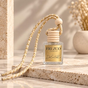 May include: A small square glass bottle with a gold label reading FREZCO CANDLES Delectable. The bottle features a ribbed wooden cap and a braided beige and gold cord with a wooden bead for hanging, designed as a car air freshener.