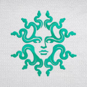 Pu&ograve; includere: Un disegno ricamato turchese di un volto con capelli a forma di serpente su uno sfondo bianco a maglia. Il volto ha un'espressione serena e i serpenti formano un motivo circolare. Il disegno &egrave; centrato sul tessuto.