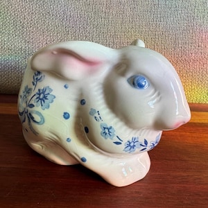 May include: A ceramic bunny figurine in a seated position. The bunny is primarily white with pink accents on the ears and nose. Blue floral patterns decorate the body, and it has blue eyes. The figurine is on a wooden surface.