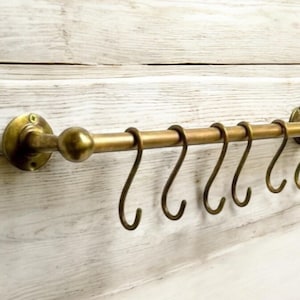 Könnte beinhalten: Ein Küchenutensilienhalter aus Messing mit acht Haken. Der Halter ist an einer weißen Wand montiert und hat einen runden Knopf am Ende.