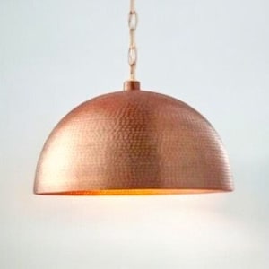 May include: A copper pendant light with a hammered finish. The light is hanging from a chain and is illuminated.