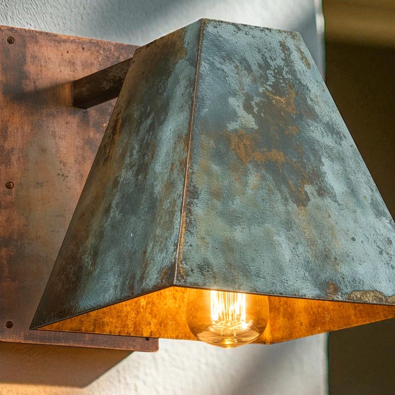 May include: A wall-mounted light fixture featuring a trapezoidal shade with a weathered, oxidized teal and copper patina finish. The interior of the shade is a warm, reflective gold tone, housing a single glowing Edison-style light bulb.