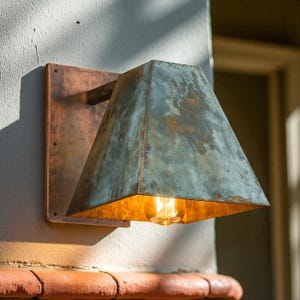 May include: A wall-mounted light fixture featuring a rectangular copper backplate and a trapezoidal shade with a weathered, oxidized teal and brown patina finish. The interior of the shade is metallic gold, housing a single glowing filament bulb.
