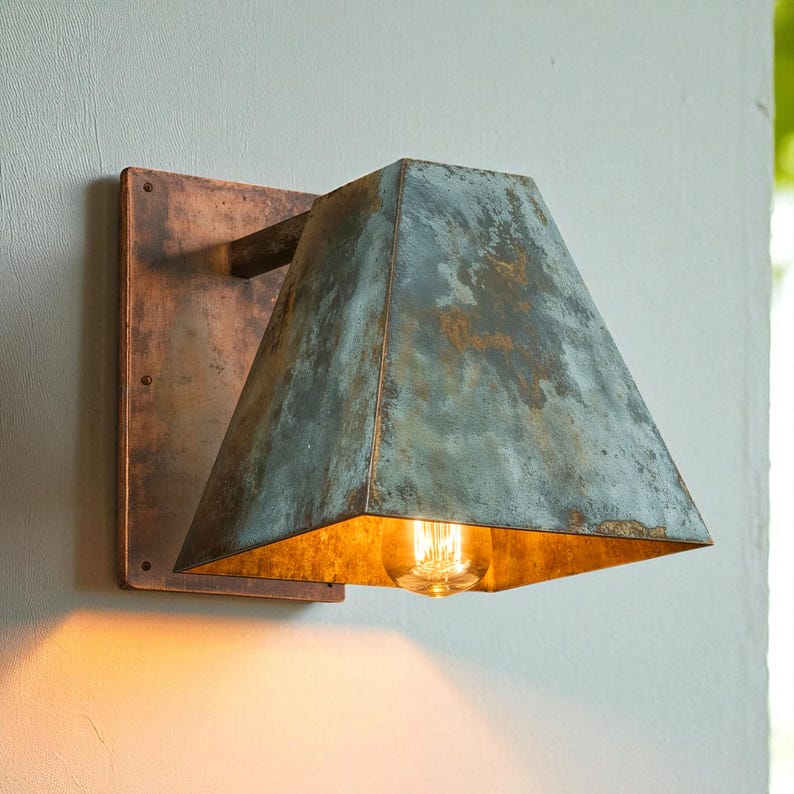 May include: A wall-mounted light fixture featuring a rectangular copper backplate and a trapezoidal shade with a weathered, oxidized blue-green patina finish. The interior of the shade is metallic, housing a single glowing light bulb.