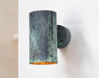 May include: A cylindrical outdoor wall sconce with a weathered, textured finish in shades of blue and green. The sconce is mounted on a circular base and emits a warm, golden light from its open bottom. The fixture is made of metal.