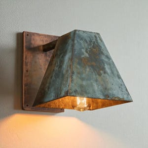 May include: A wall-mounted light fixture featuring a rectangular copper backplate and a trapezoidal shade with a weathered, oxidized teal and brown patina finish. The interior of the shade houses a single exposed light bulb emitting a warm glow.