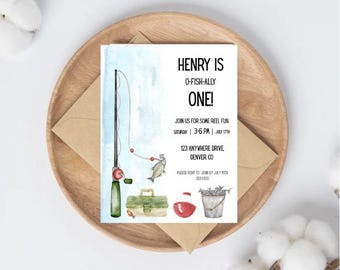 Peut inclure: Une invitation à thème pêche à l'aquarelle. L'invitation présente des illustrations de canne à pêche, de boîte à pêche et de poissons. Le texte sur l'invitation se lit « HENRY IS O-FISH-ALLY ONE ! » et comprend les détails de l'événement. L'invitation est posée sur une assiette en bois avec une enveloppe.