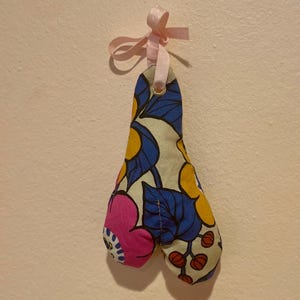 May include: A fabric decoration with a floral design in blue, yellow, and pink. The ornament is shaped like a teardrop with two rounded sections at the bottom. It hangs from a pink ribbon tied in a bow.