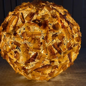 Könnte beinhalten: Eine kugelförmige Lampe mit warmem Glanz. Die Lampe besteht aus verflochtenen Strängen und strukturiertem, hellbraunem, organischem Material, wodurch eine einzigartige Oberfläche entsteht. Die interne Lichtquelle wirft ein weiches, Umgebungslicht.