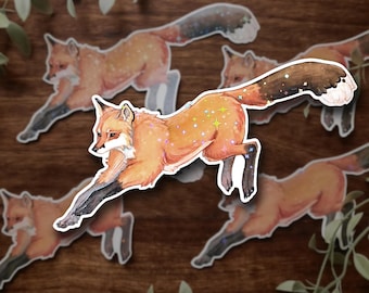 May include: A collection of stickers featuring watercolor illustrations of foxes in motion. The foxes are depicted in shades of orange, brown, and white, with sparkling accents on their bodies and tails. The stickers are on a wooden background.