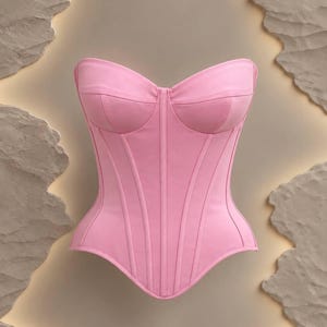 May include: A strapless corset top in a solid light pink color. The garment features a sweetheart neckline, structured vertical paneling throughout the bodice, and a pointed hemline that dips at the center front and rises at the sides.