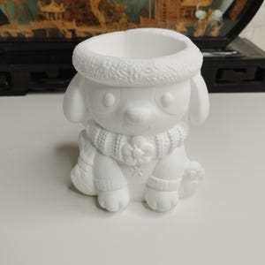 May include: A white ceramic puppy-shaped planter. The puppy is wearing a scarf and a hat. The planter has a round opening at the top for plants or other items. The design is detailed and textured.